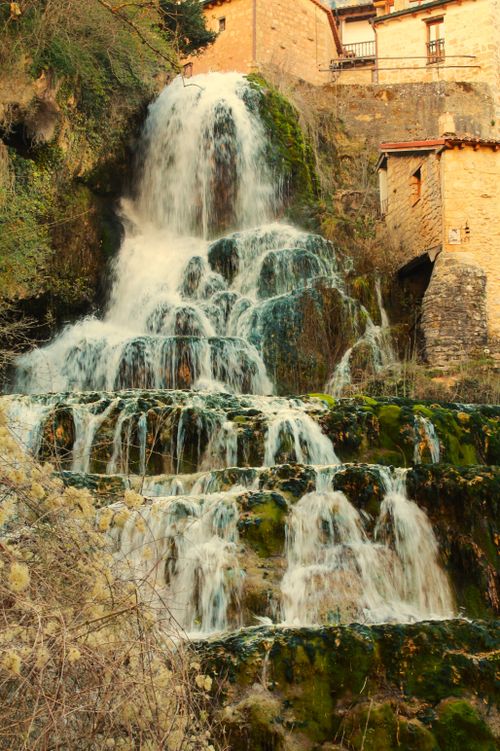 Free Scenic waterfall cascades amidst rocky terrain in a quaint town setting, highlighting nature and architecture. Stock Photo