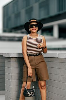 Fashionable woman in sunglasses and hat posing against urban backdrop.