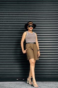 Stylish woman posing in shorts and hat against a gray wall.
