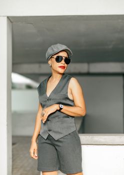 Portrait of a woman in gray vest and shorts, wearing sunglasses and a cap for urban fashion look.
