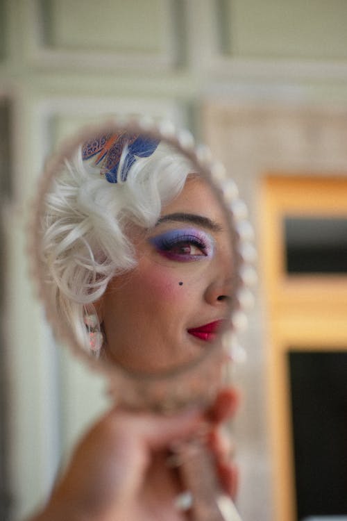 A woman with white makeup and purple hair is looking in a mirror