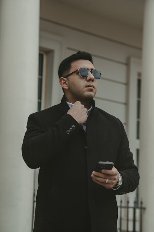 A man in a black coat and sunglasses looking at his phone
