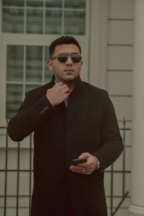 A man in a black coat and sunglasses is looking at his phone