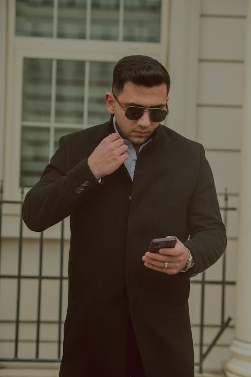 A man in a black coat and sunglasses looking at his cell phone