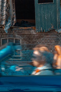 A motion blur captures a person driving a blue convertible past an aged brick structure with a dilapidated window.