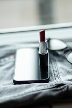A close-up of red lipstick standing on a smartphone with eyeglasses, suggesting elegance and modern lifestyle.