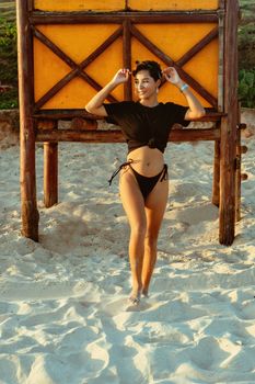 Woman in black bikini posing by a lifeguard stand on a sunny beach.