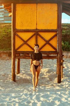 Woman in bikini standing on sandy beach under wooden structure at sunset.
