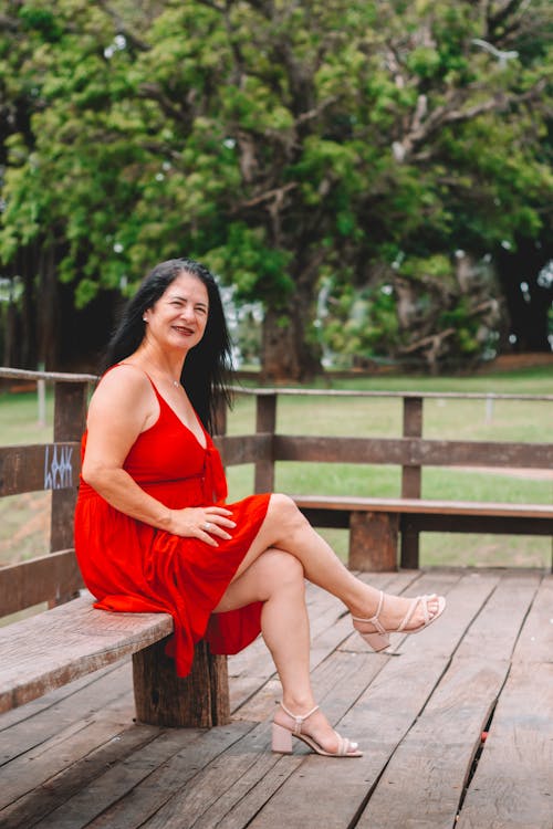 Happy Brunette Woman in Red Dress Sitting on Wooden Bench in Park