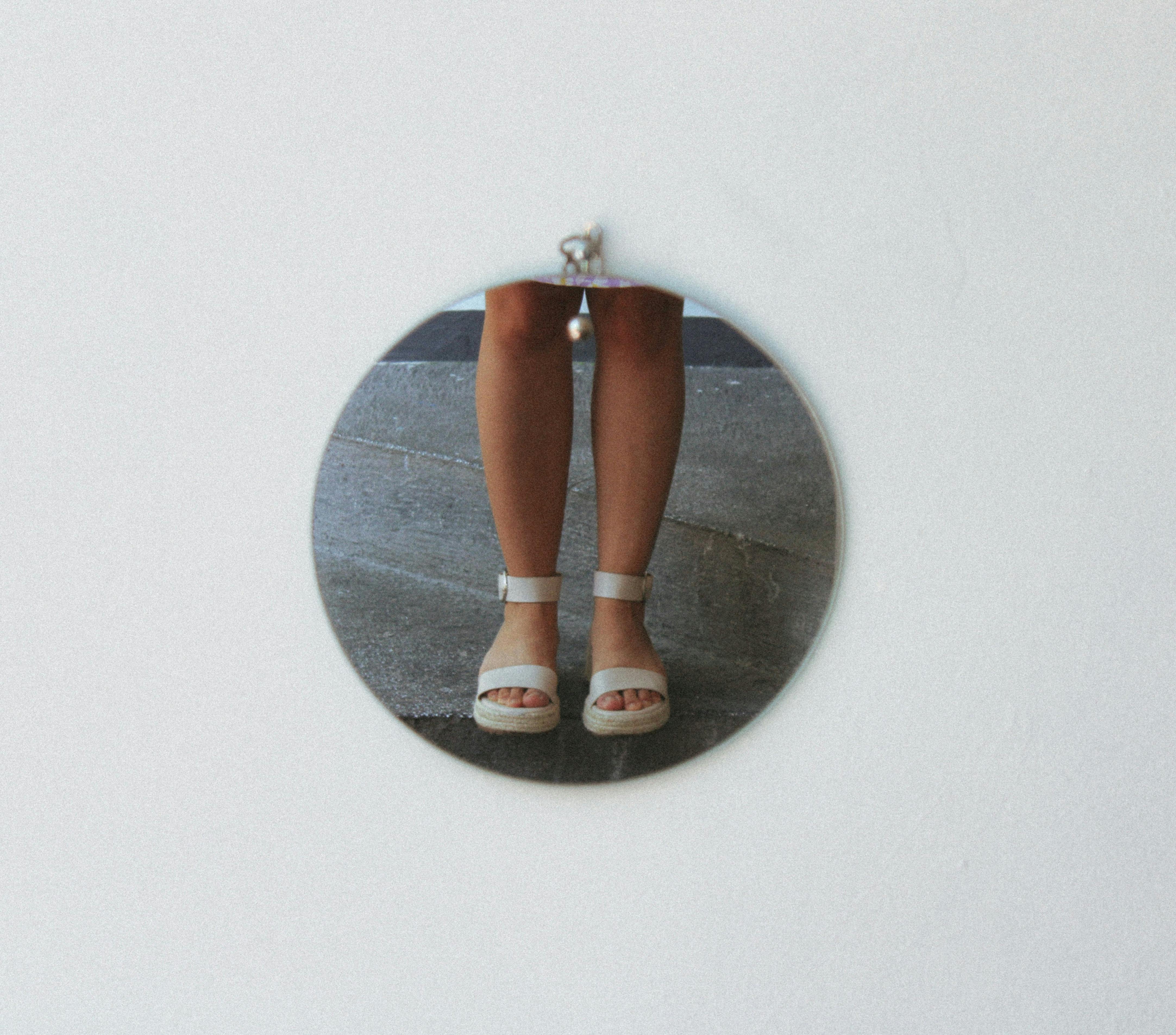 Close-up of child's legs and sandals reflected in a round mirror on a white wall.