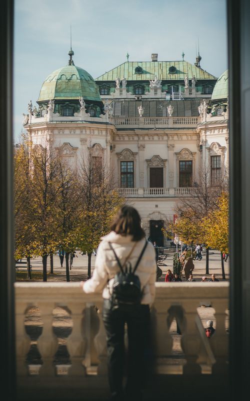 Free A woman stands on a balcony viewing Belvedere Palace surrounded by autumn trees in Vienna. Stock Photo
