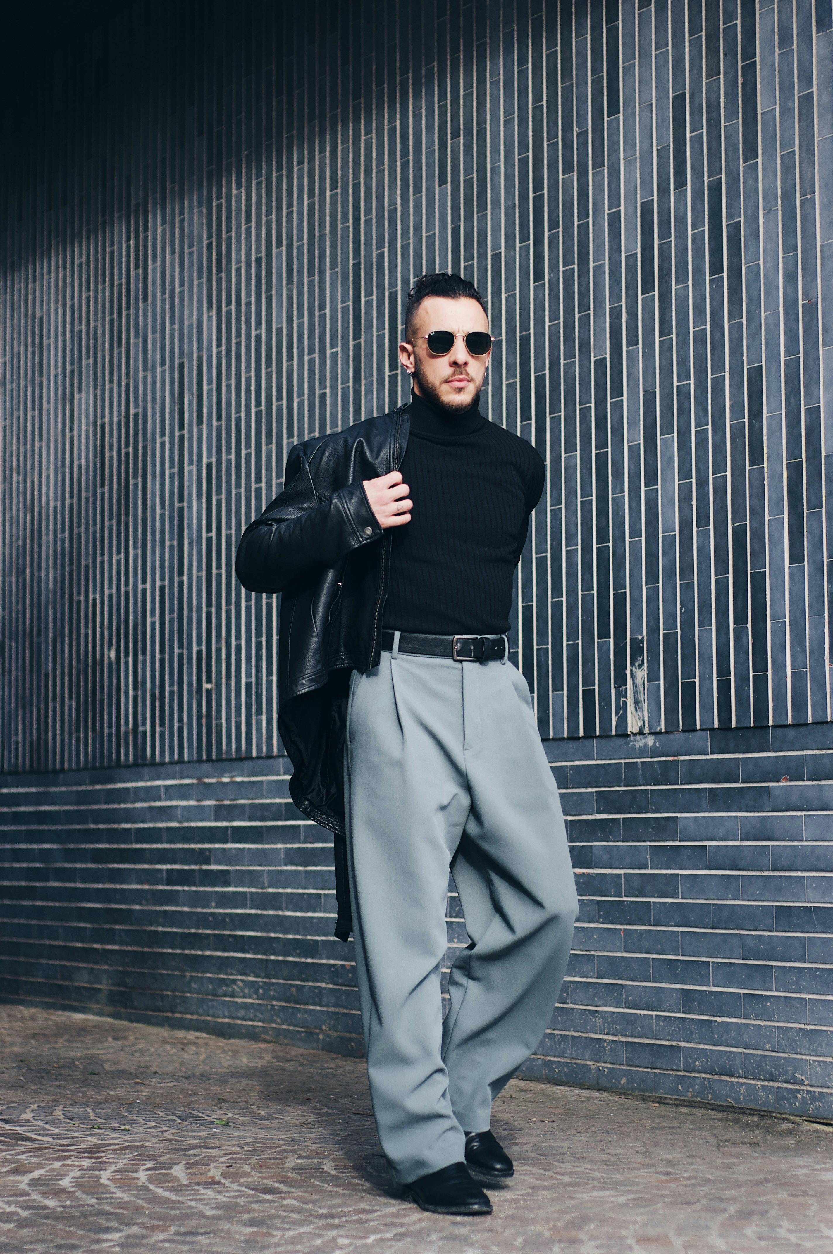 Fashionable man in Italy wearing sunglasses, leather jacket, and suit pants against a tiled wall.
