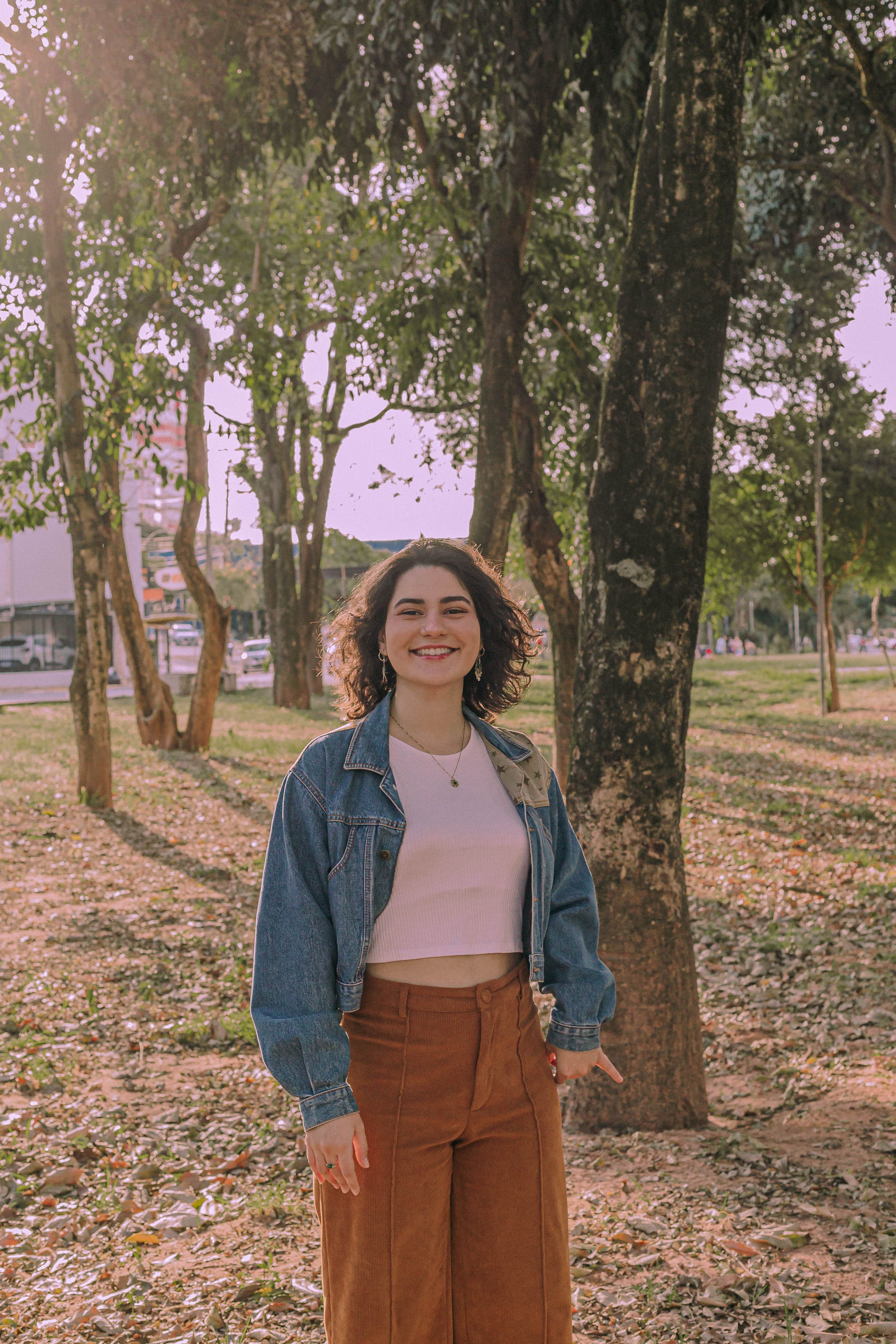 A woman in a jean jacket smiles warmly while standing in a sunlit park in Presidente Prudente, Brazil.