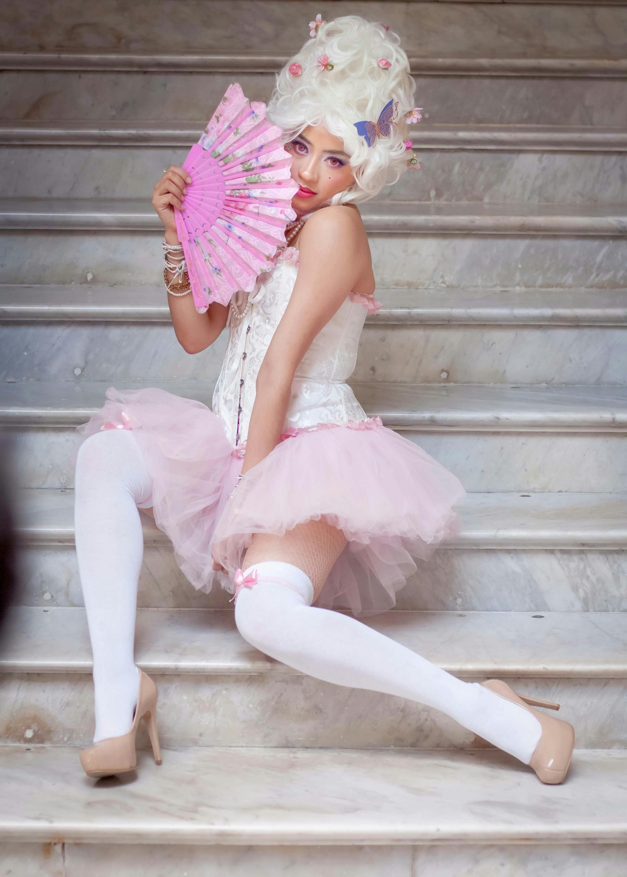 Model in Victorian-inspired outfit sitting on marble stairs with pink fan.