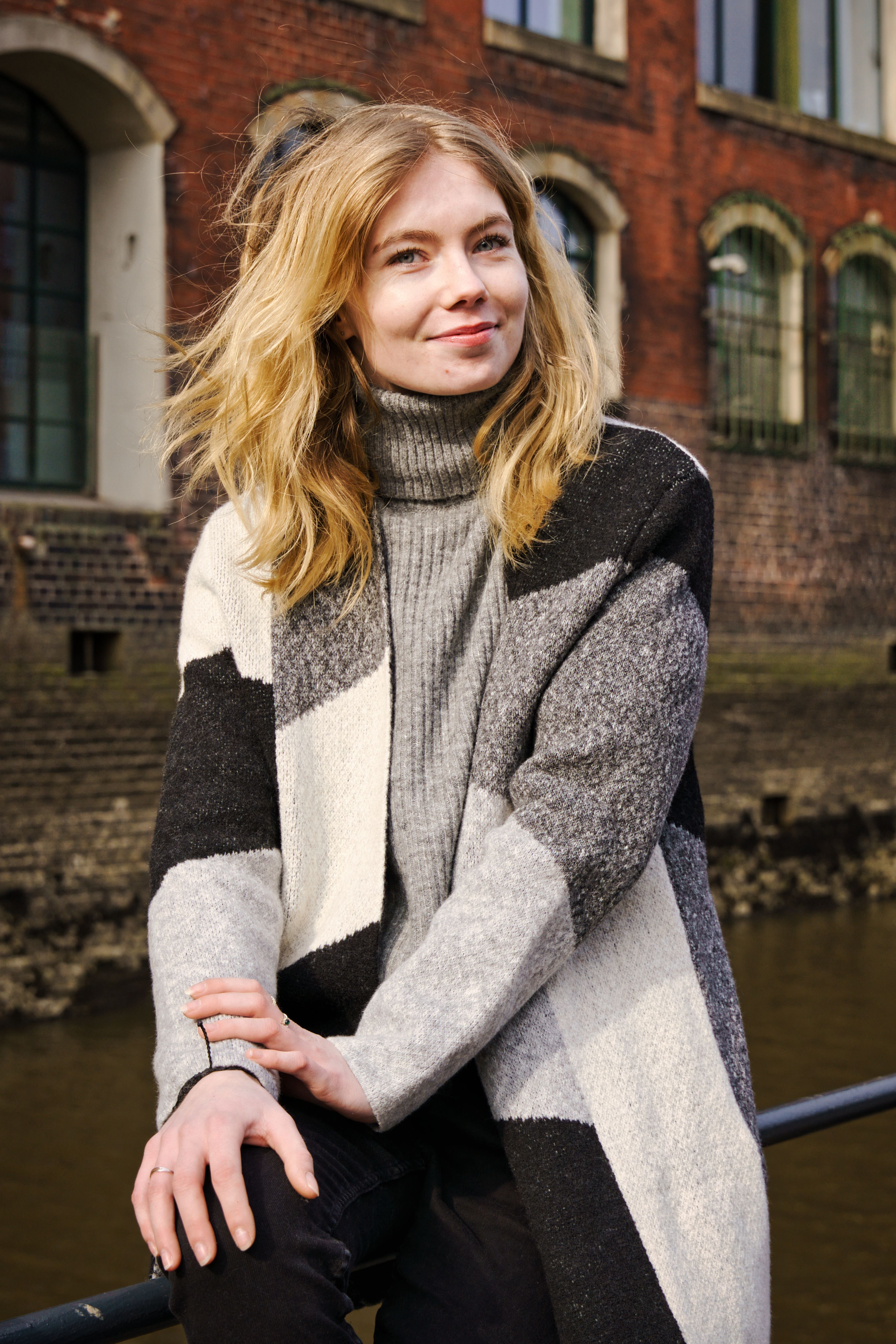 Portrait of a smiling woman sitting by a canal in Hamburg, Germany, wearing a fashionable sweater.