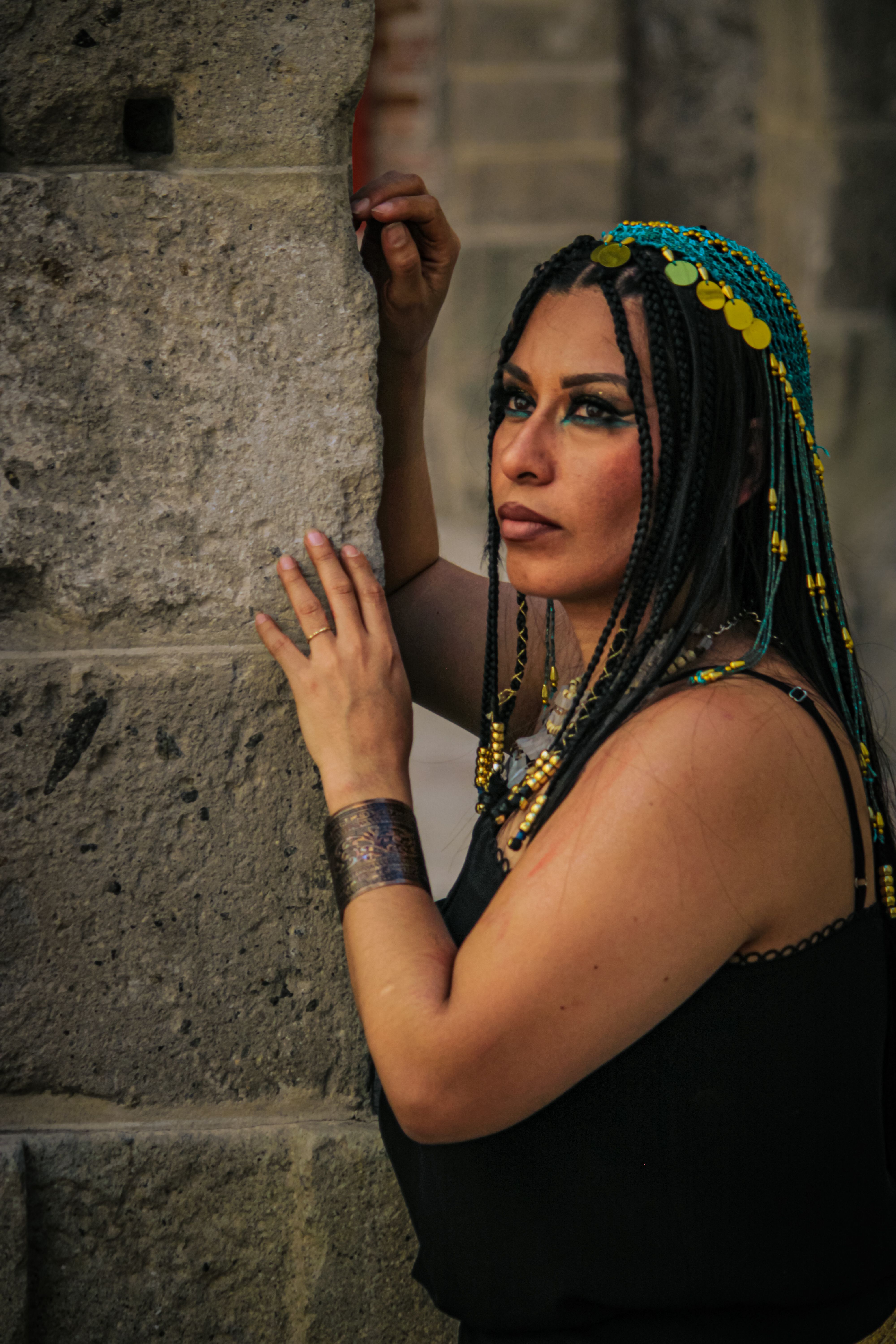 Woman with braided hair and vibrant makeup poses by a historic stone wall in daylight.