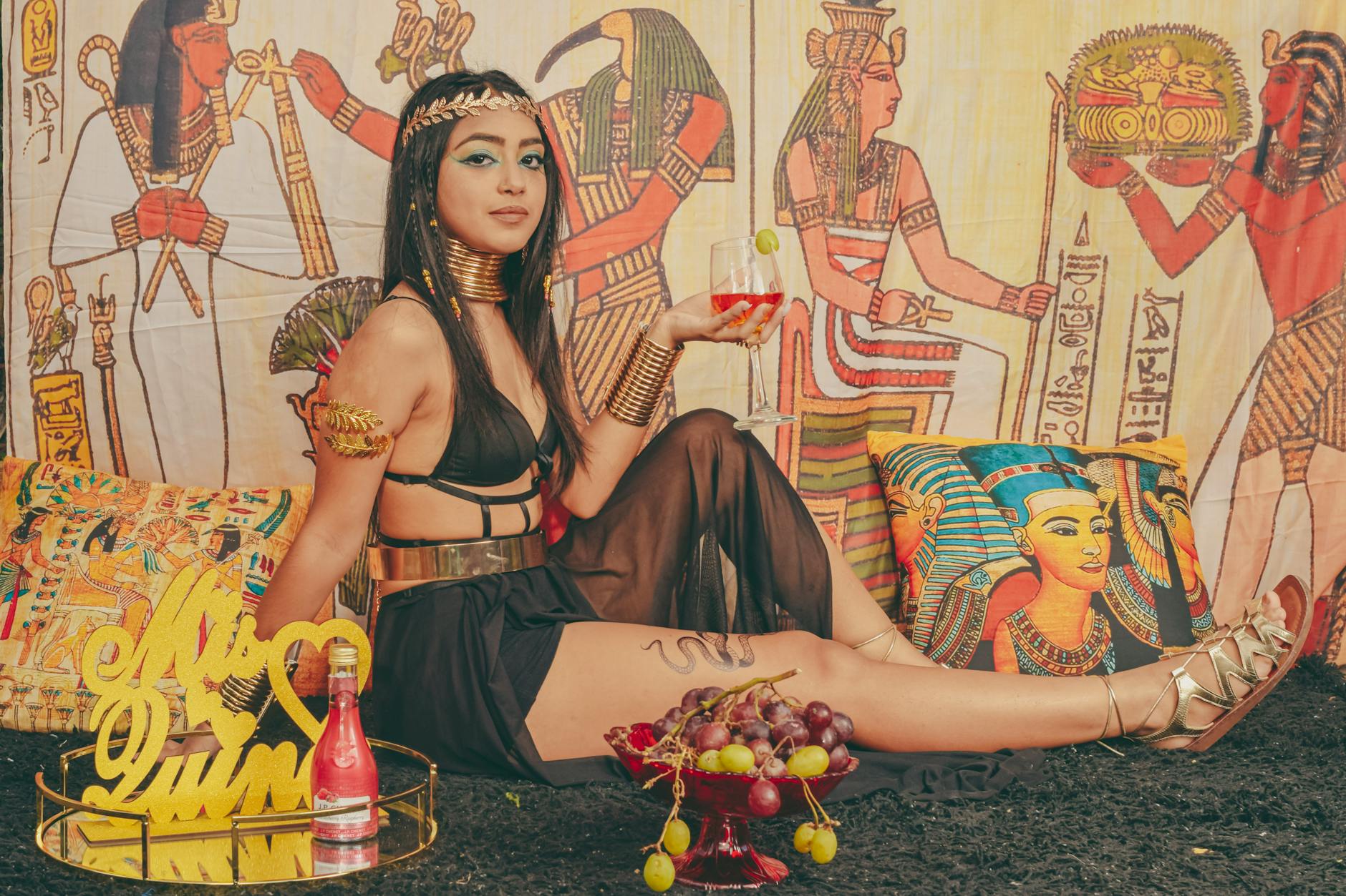 Stylized photo of a model in Egyptian-themed attire, holding a drink indoors.