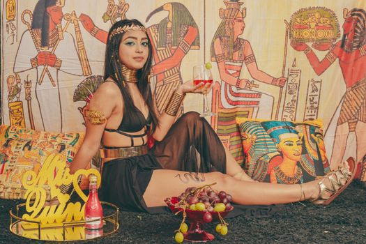 Stylized photo of a model in Egyptian-themed attire, holding a drink indoors.
