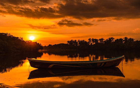 Silhouetted boat on a tranquil lake with vibrant orange sunset reflecting on water.