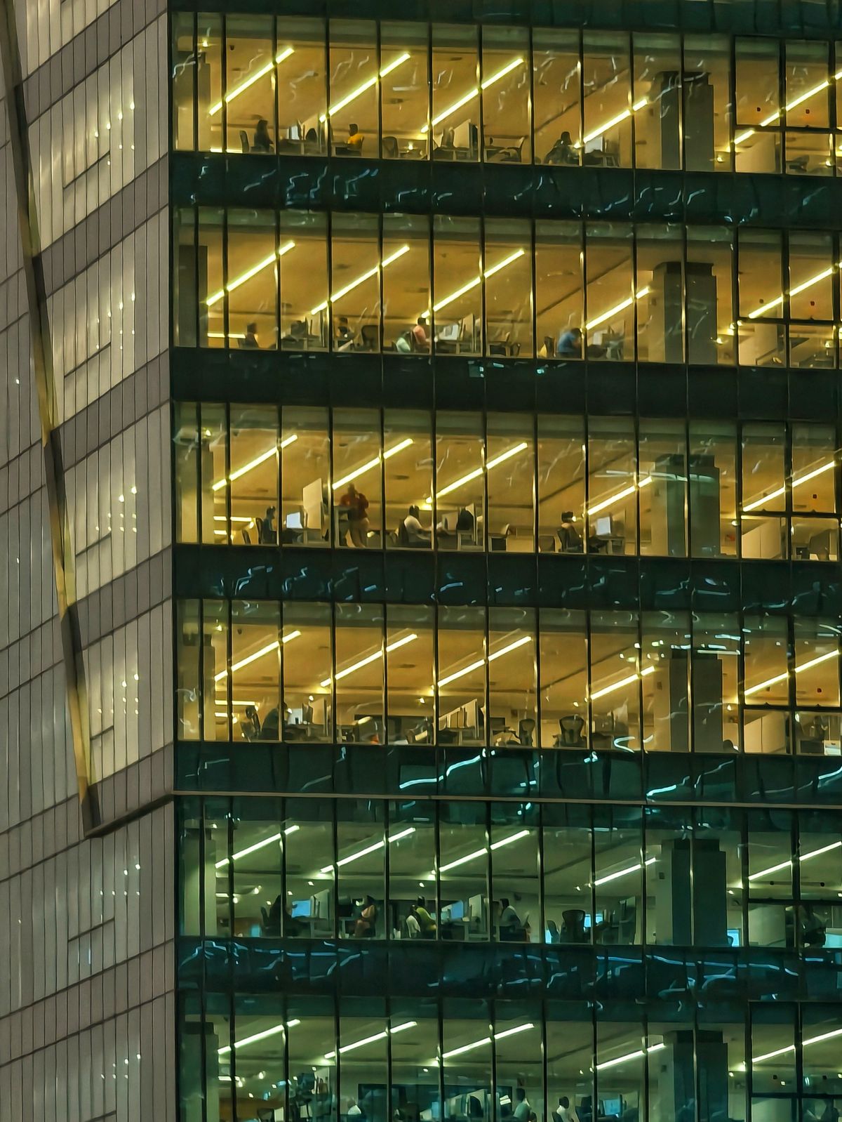 Rows of lit floors in an office building at night, with people still at their desks long after the workday ended, work continuing in a building nobody is watching