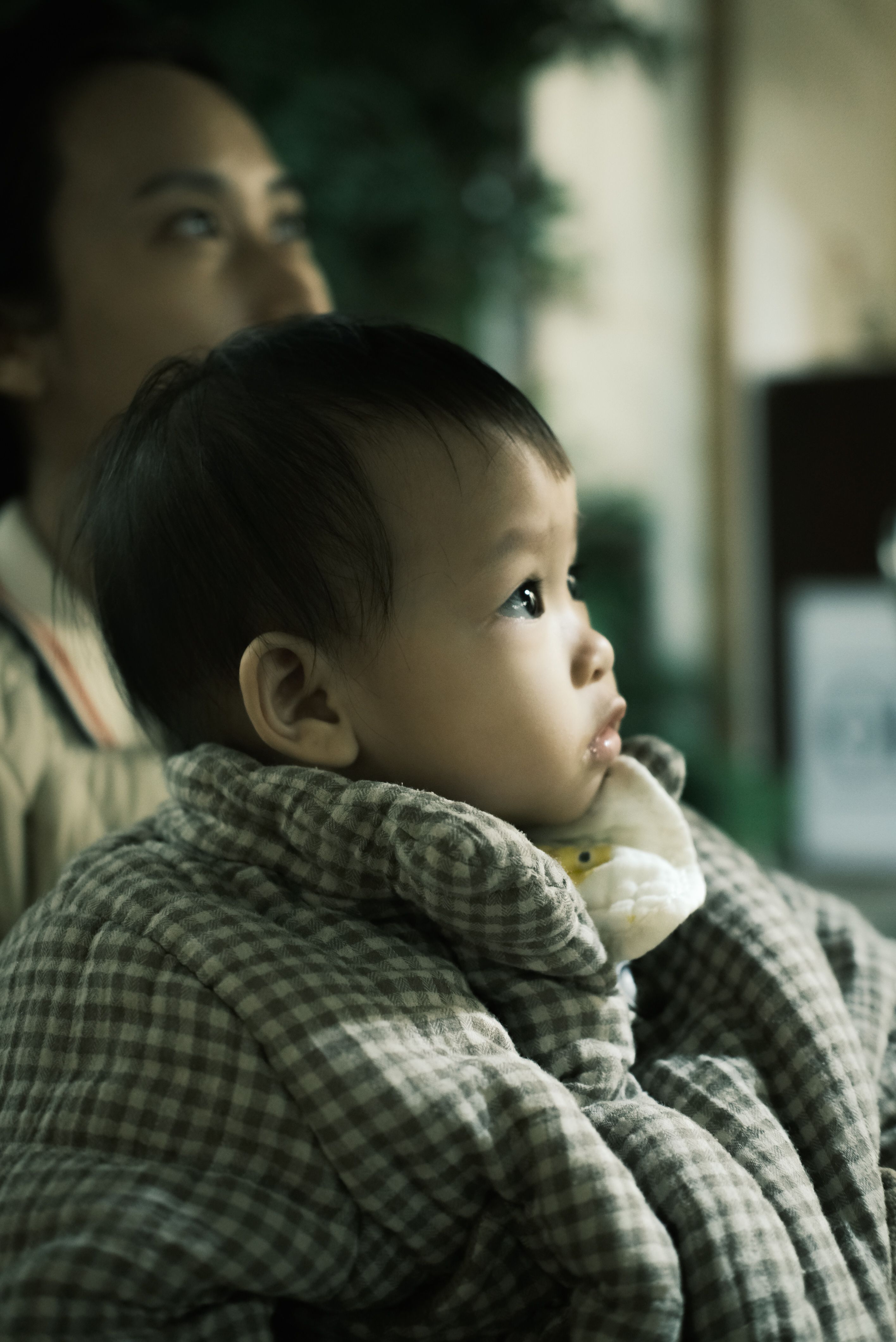 A baby in warm clothing gazes intently indoors, accompanied by an adult.