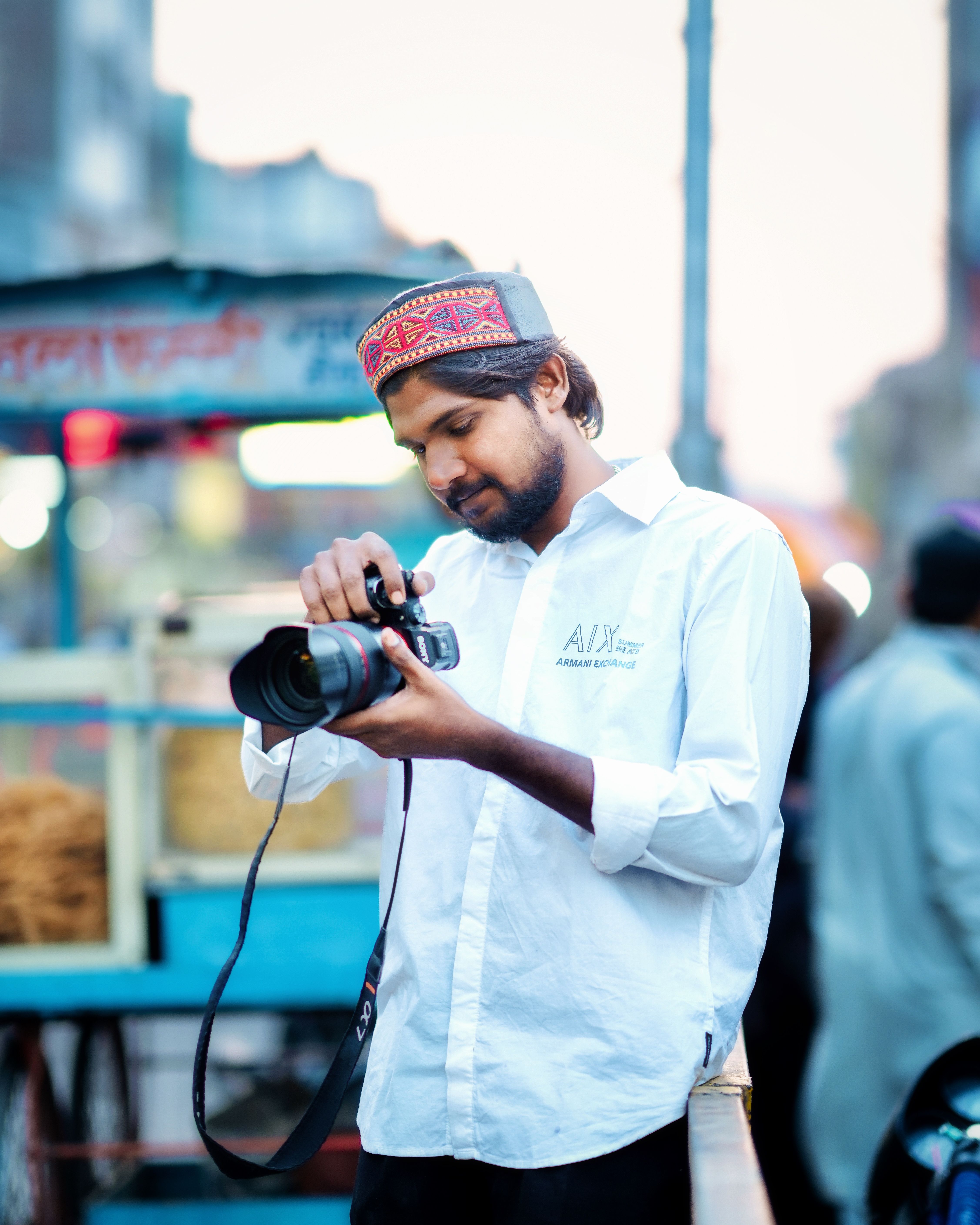 South Asian man in traditional cap capturing street scenes with DSLR.