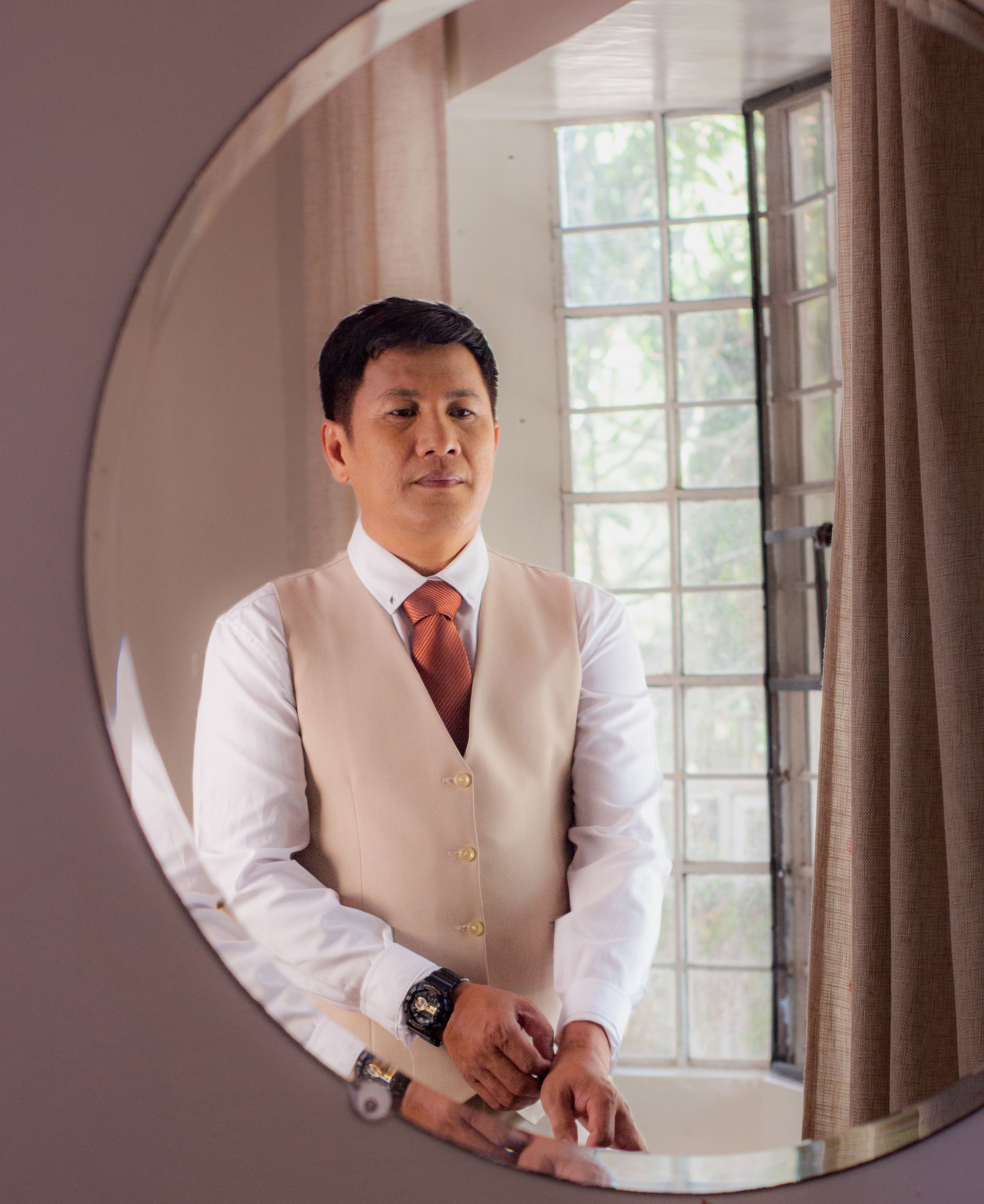 An Asian man in formal attire adjusts his tie in front of a mirror indoors, with natural light.