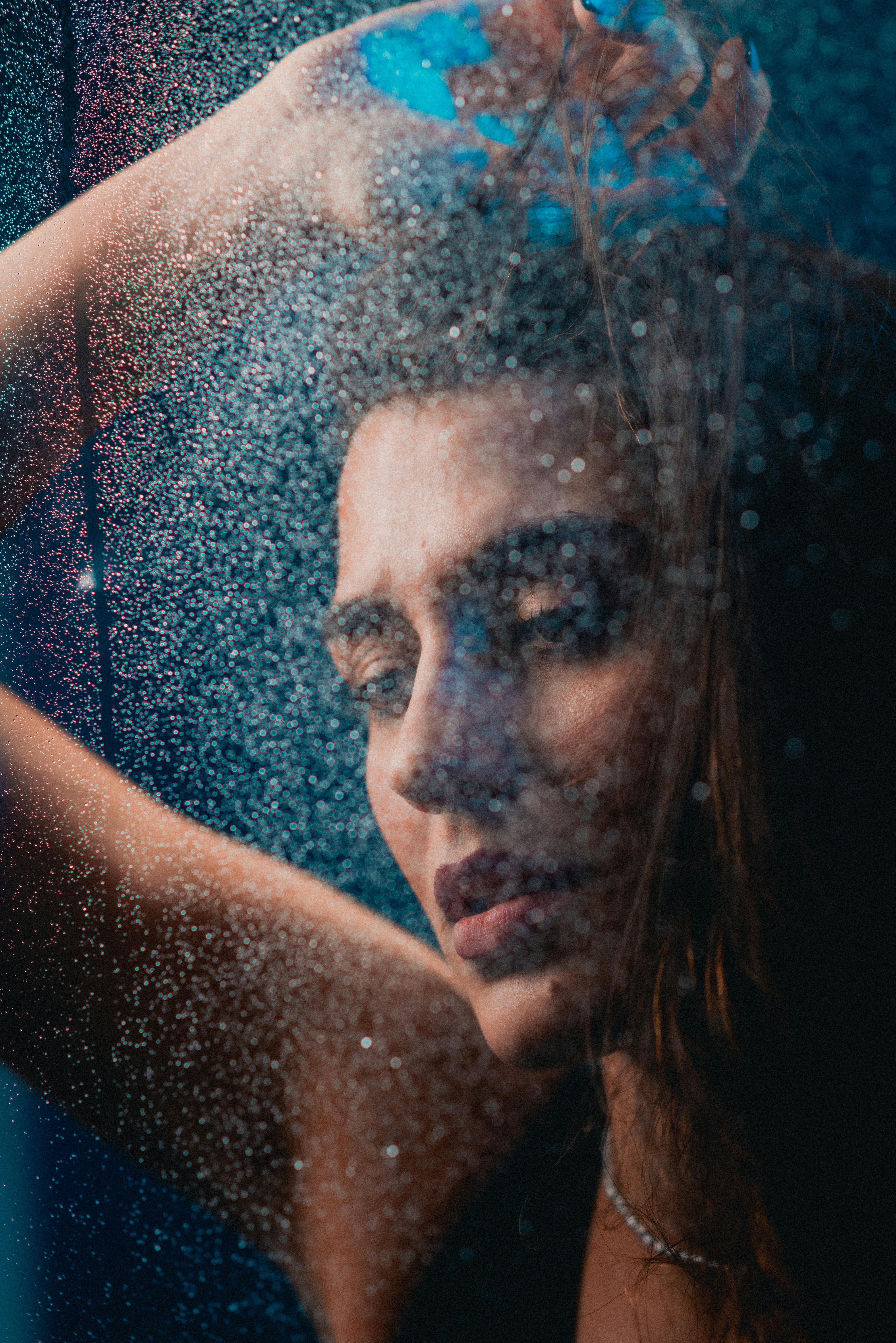 Emotive portrait of a woman with raised arm behind rainy glass, creating a contemplative mood.