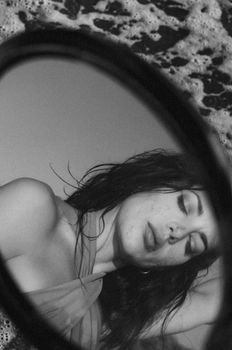 Black and white portrait of a woman reflected in a mirror beside the sea.