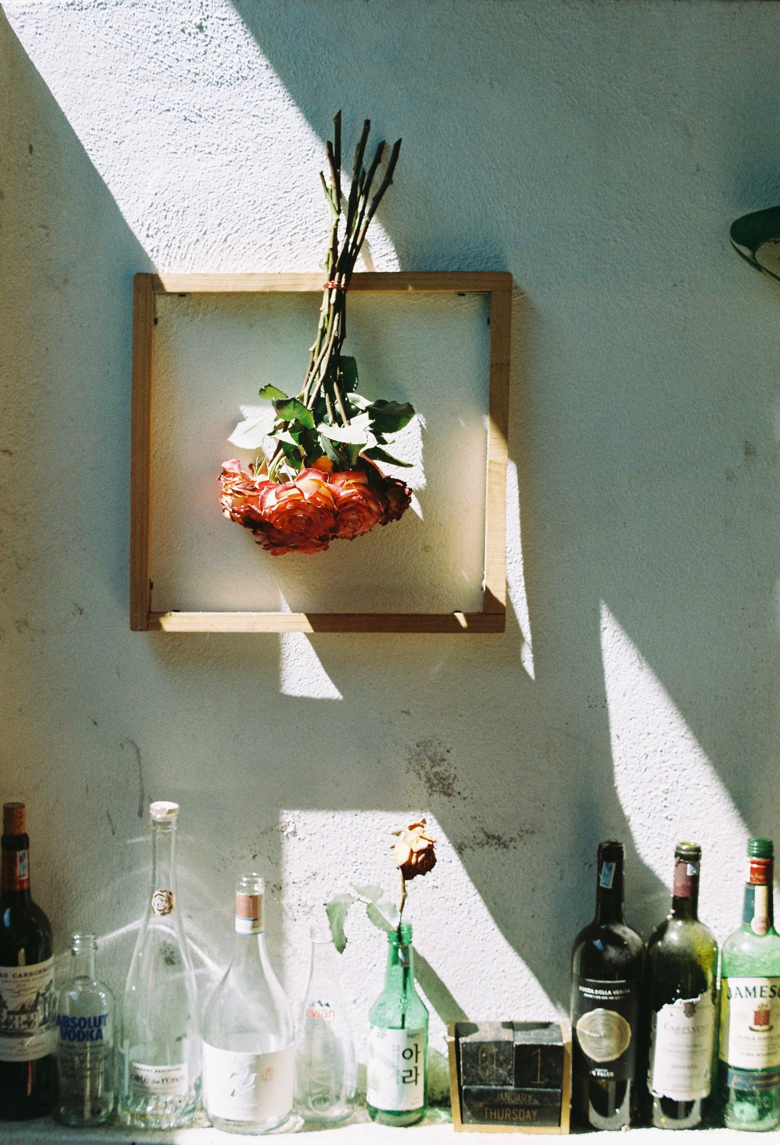 Framed upside down roses above a collection of wine bottles on a sunlit wall.