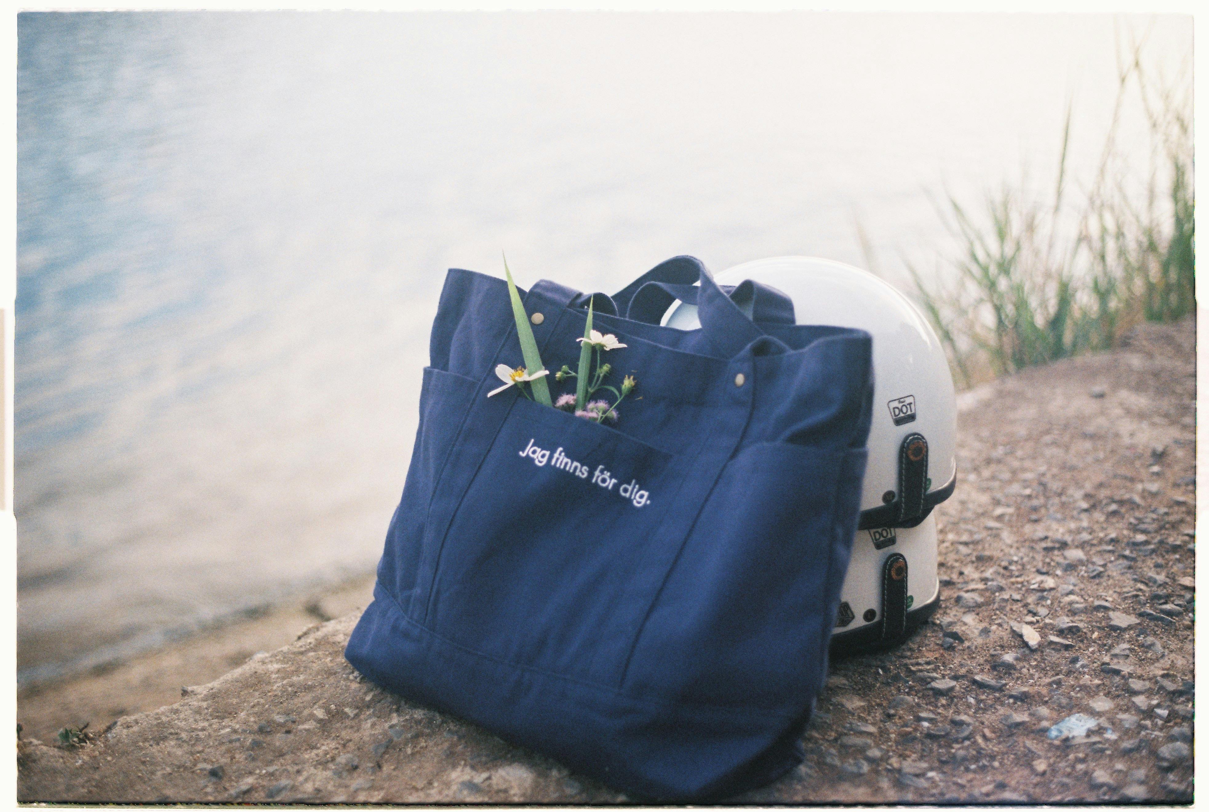 A blue tote bag with wildflowers and a helmet by the tranquil riverbank.
