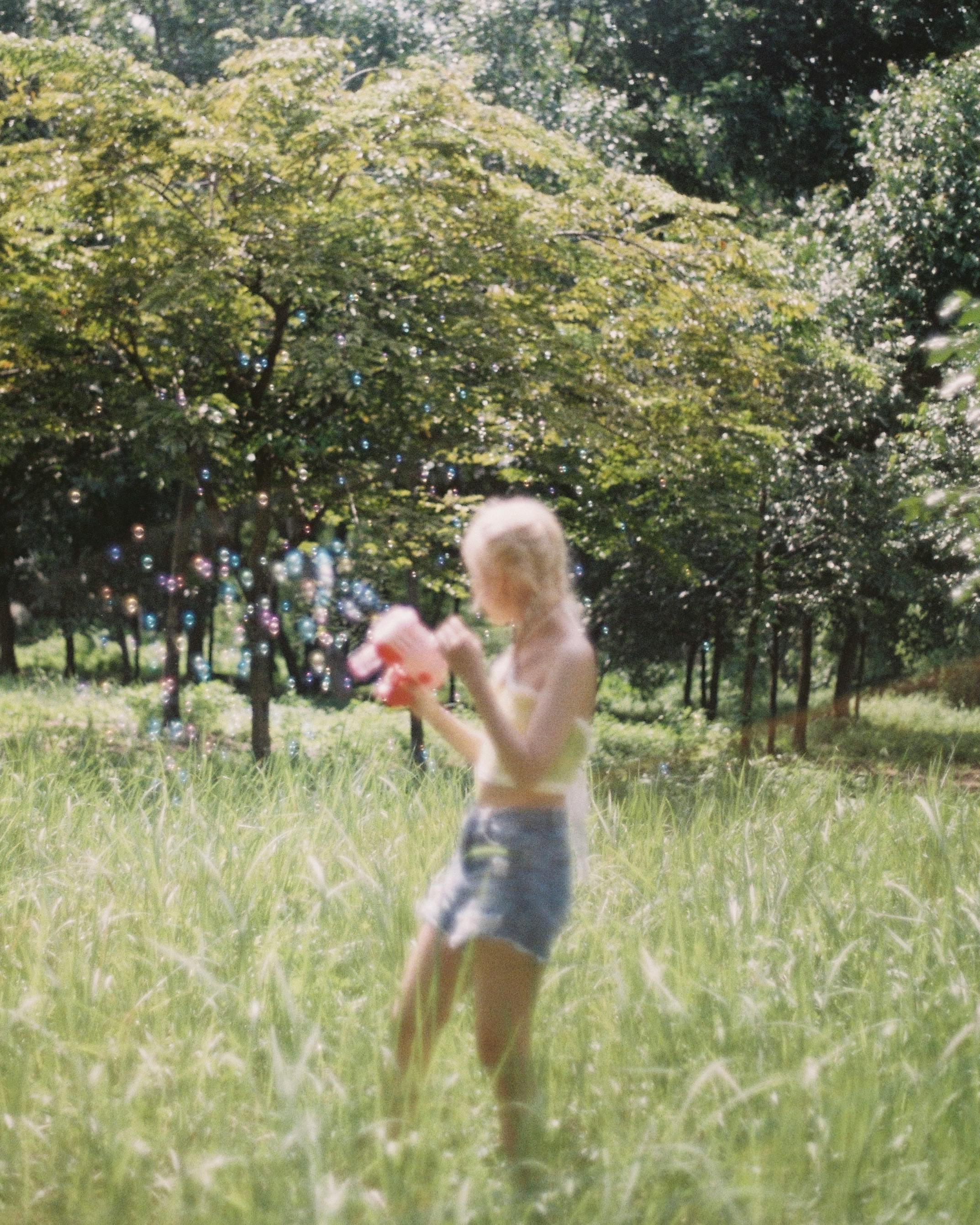 Blonde woman joyfully blowing bubbles in a sunny meadow surrounded by lush greenery.