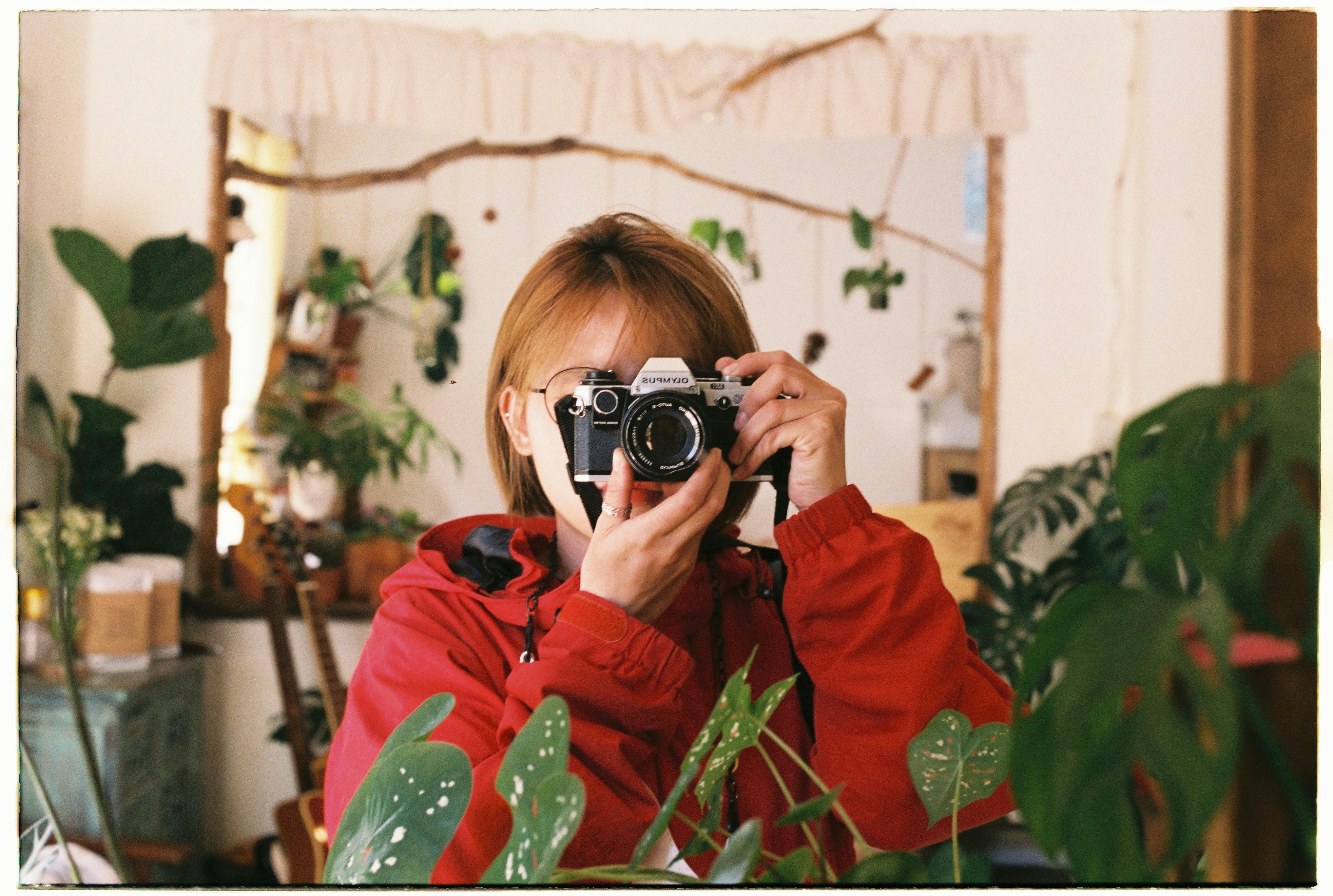 A woman in a red jacket captures the moment with an analogue camera amidst indoor plants.