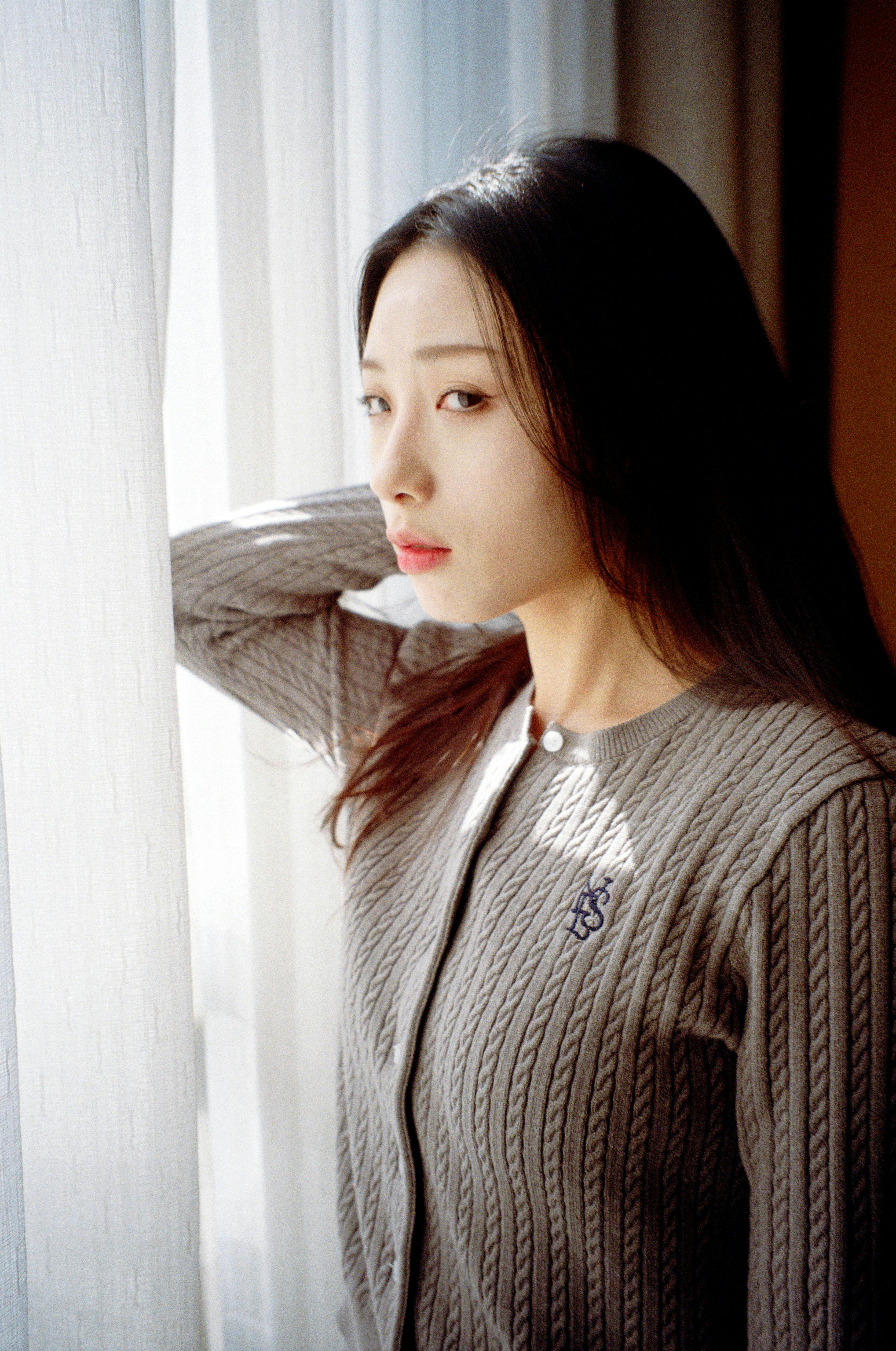 A serene portrait of a fashionable woman standing beside a sunlit window indoors.