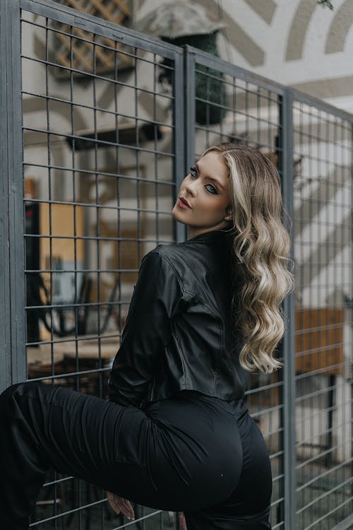 A woman in black pants and jacket posing in front of a fence