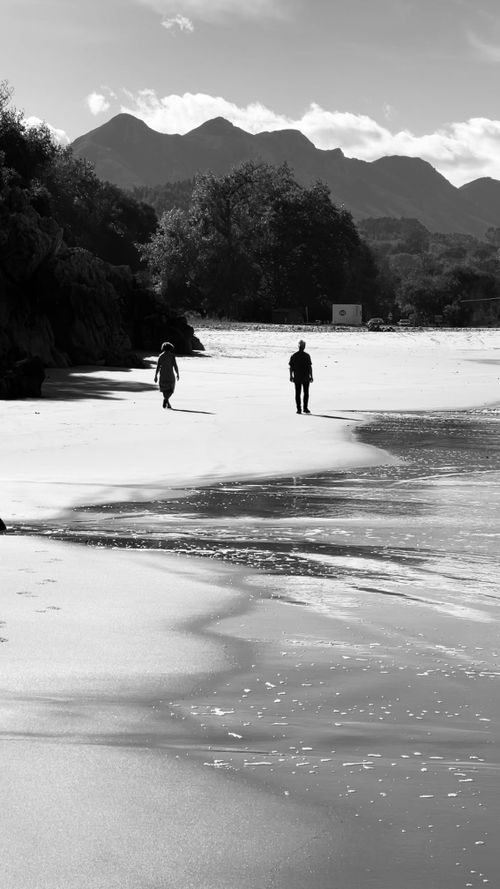 Free Two people walking on a tranquil beach against a mountainous backdrop. A peaceful, monochromatic scene. Stock Photo