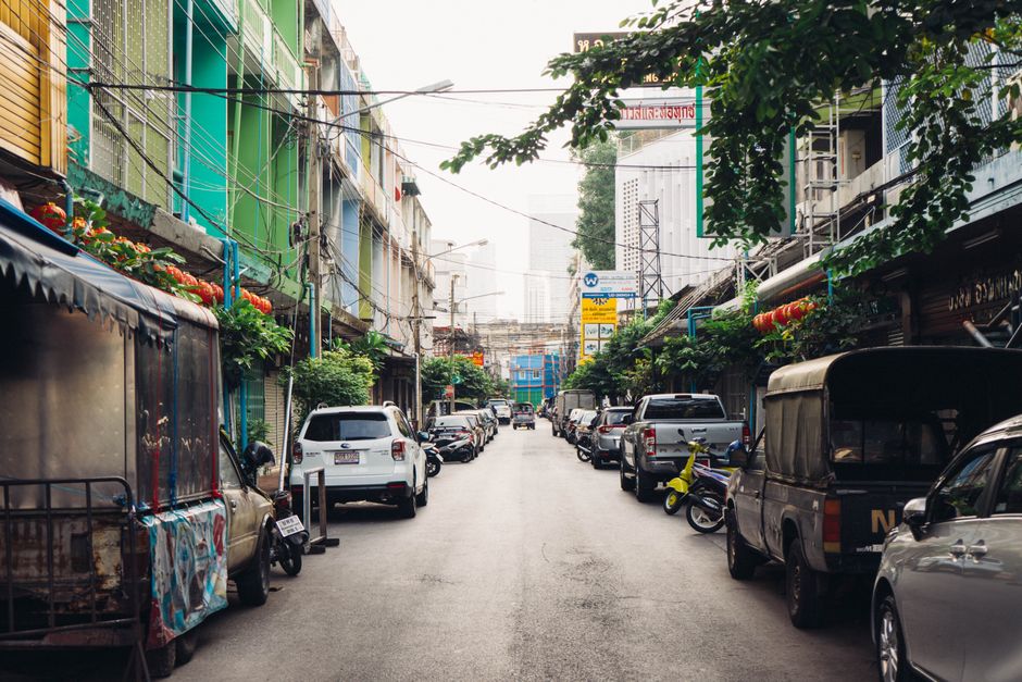 Bustling street in Bangkok with colorful buildings and parked vehicles on a sunny day.