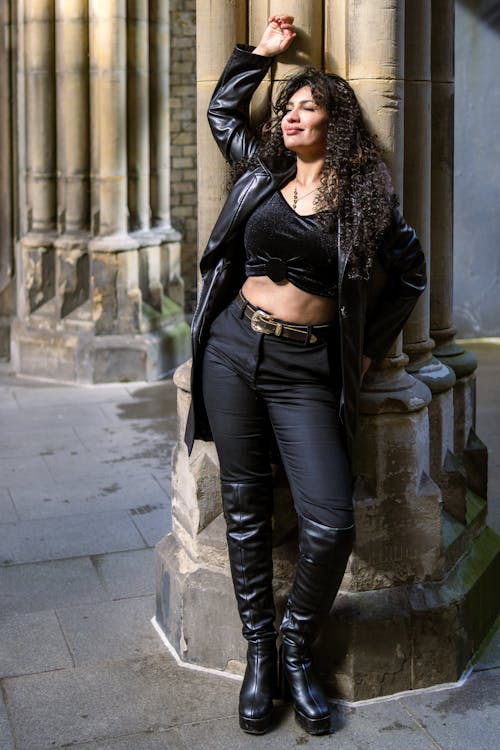 A woman in black leather pants and boots leaning against a pillar