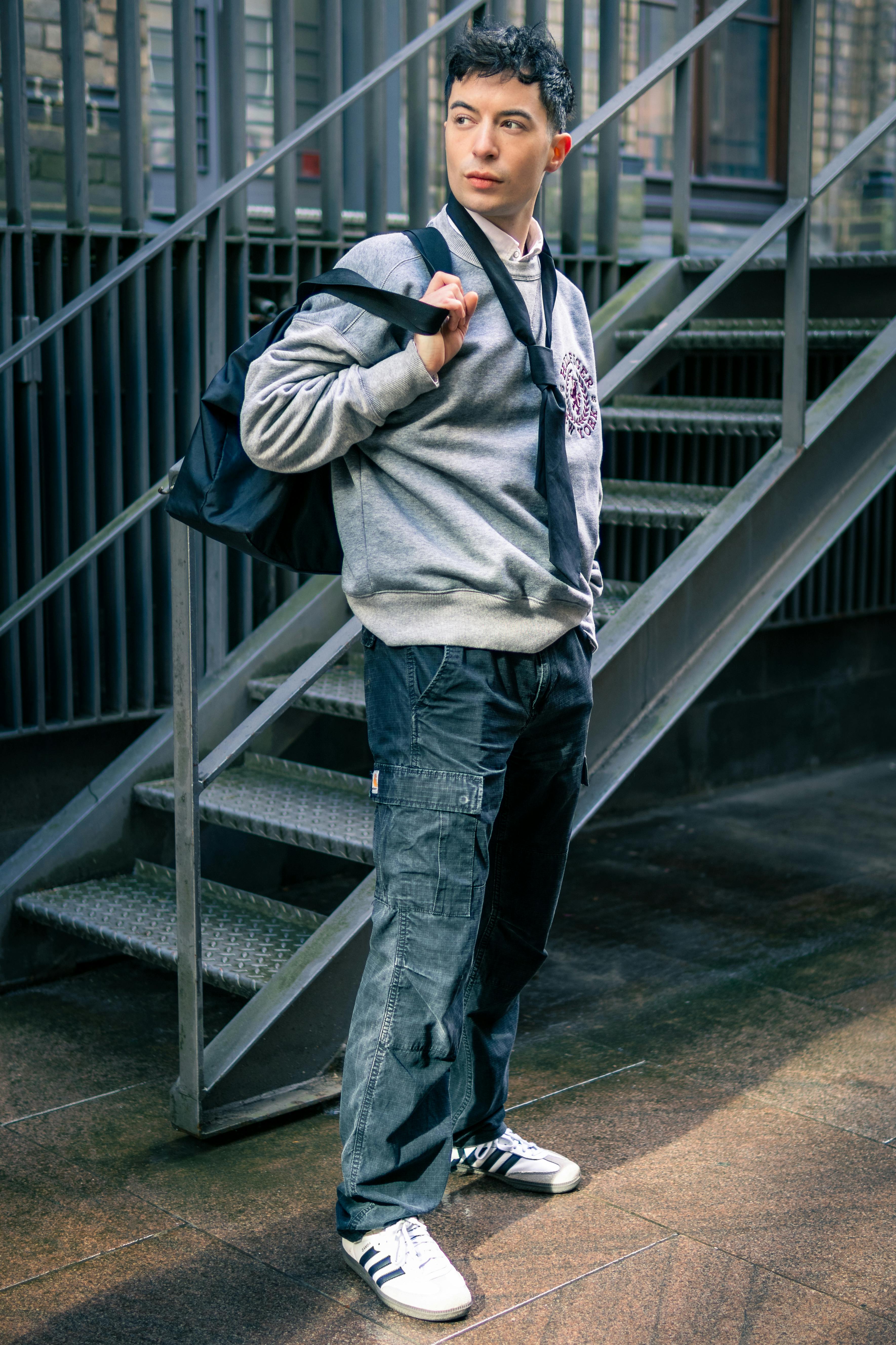 Young man in casual fashion stands by an outdoor staircase, holding a bag confidently.