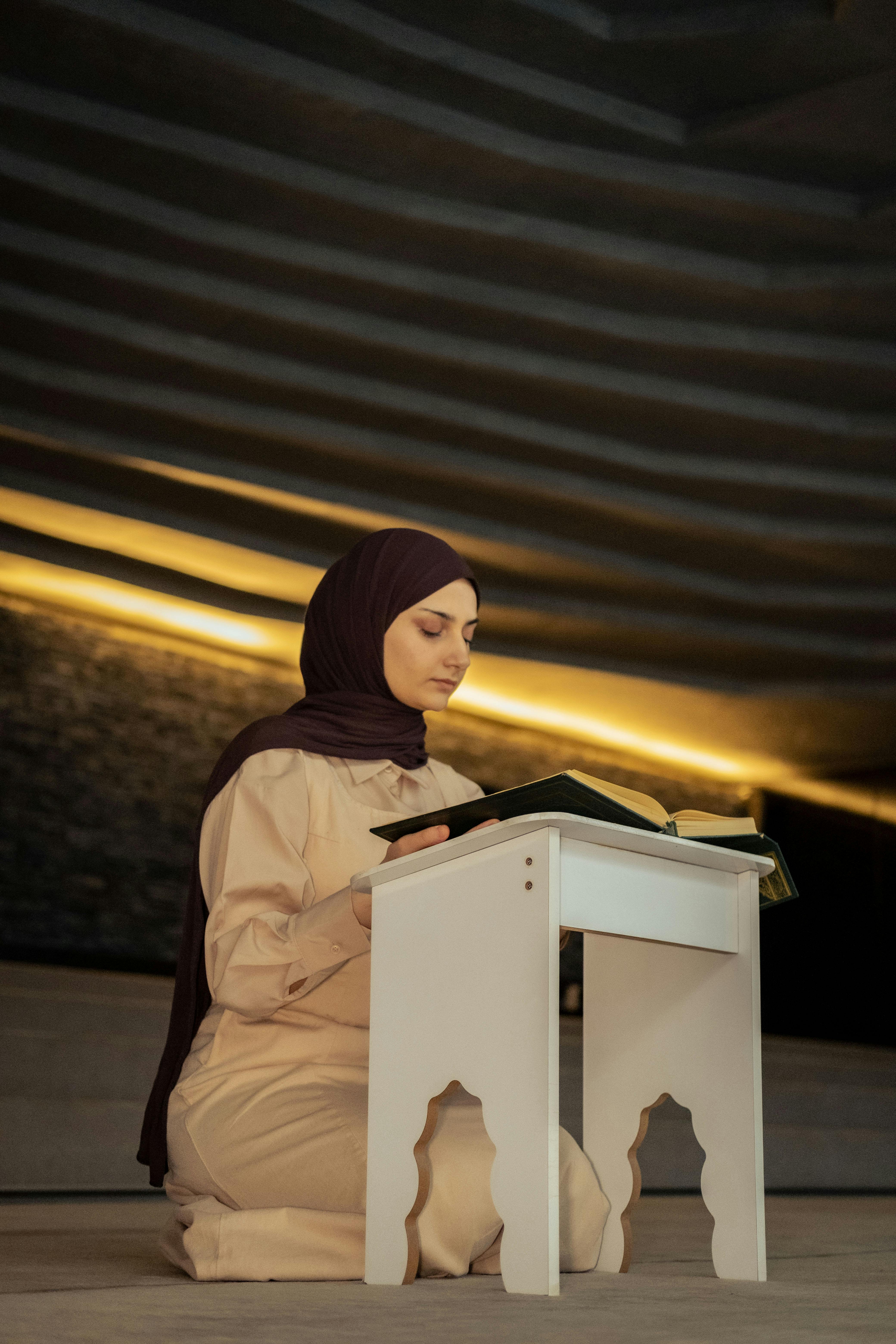 A woman in a headscarf kneeling indoors, deeply engaged in reading and prayer.