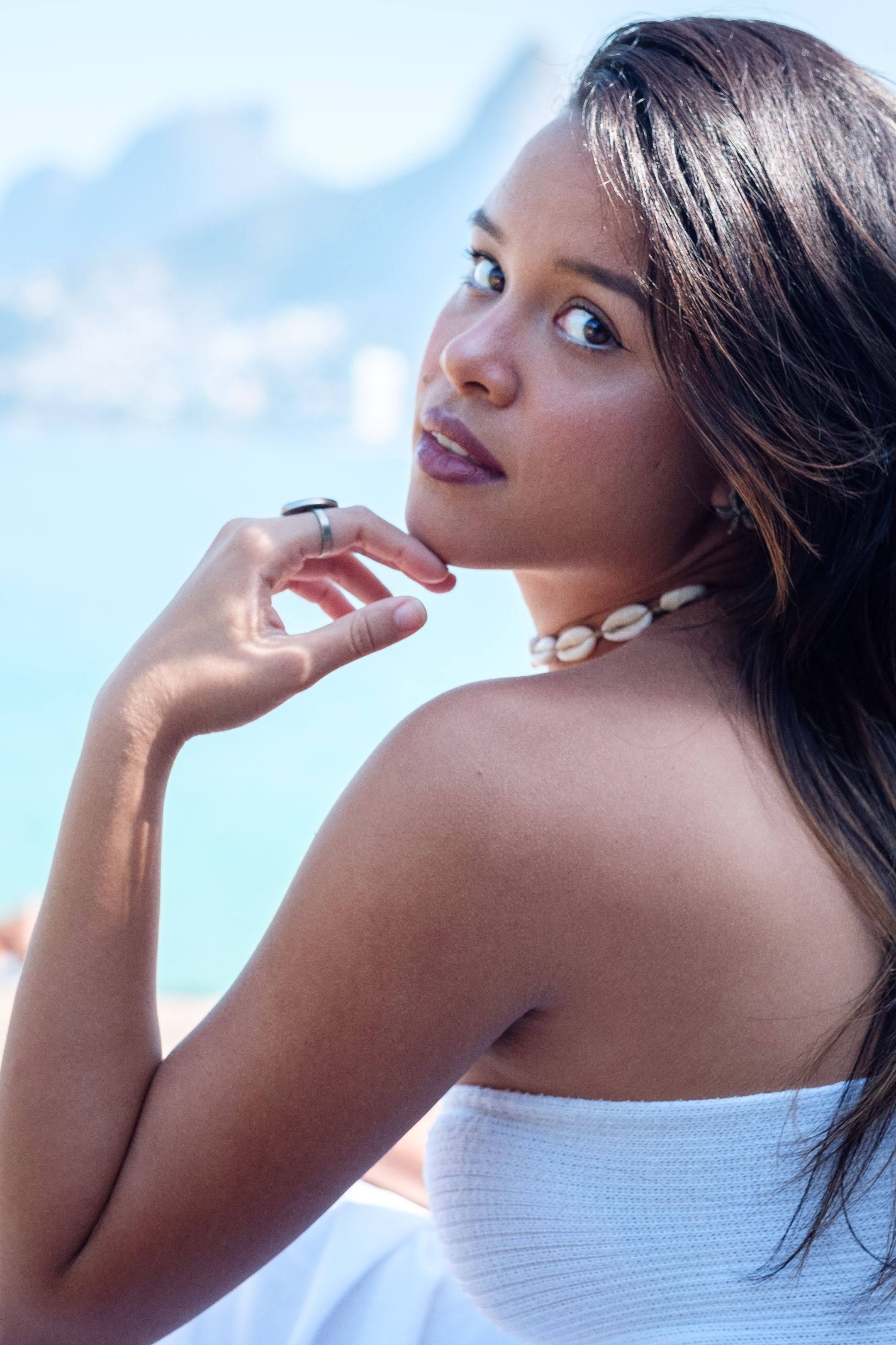 Elegant Brazilian woman in white dress poses gracefully with ocean backdrop on a sunny day.