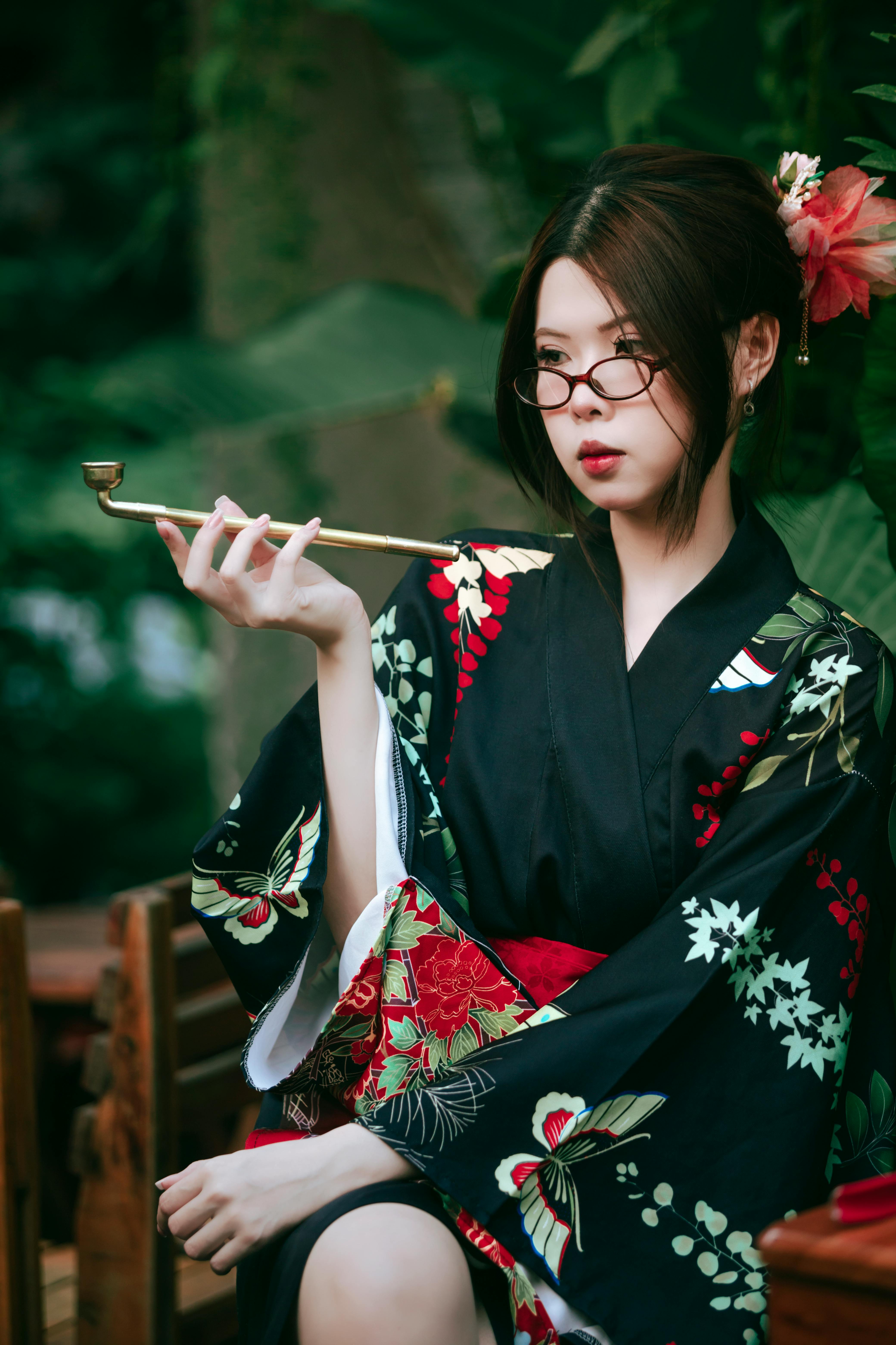 A young woman wearing a floral patterned kimono poses gracefully with glasses in a lush garden setti