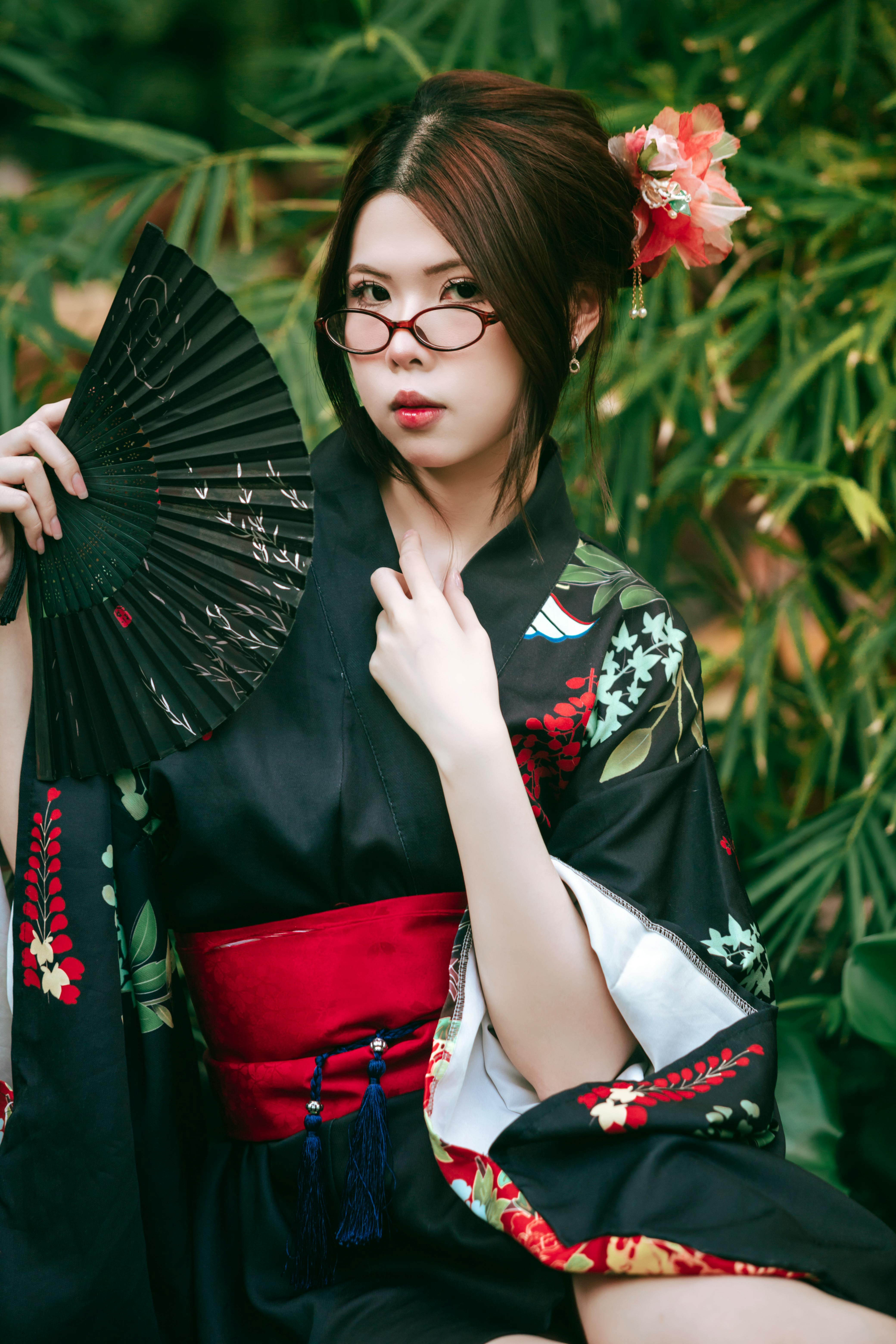 An elegant portrait of a woman in a traditional kimono holding a fan amidst lush greenery.