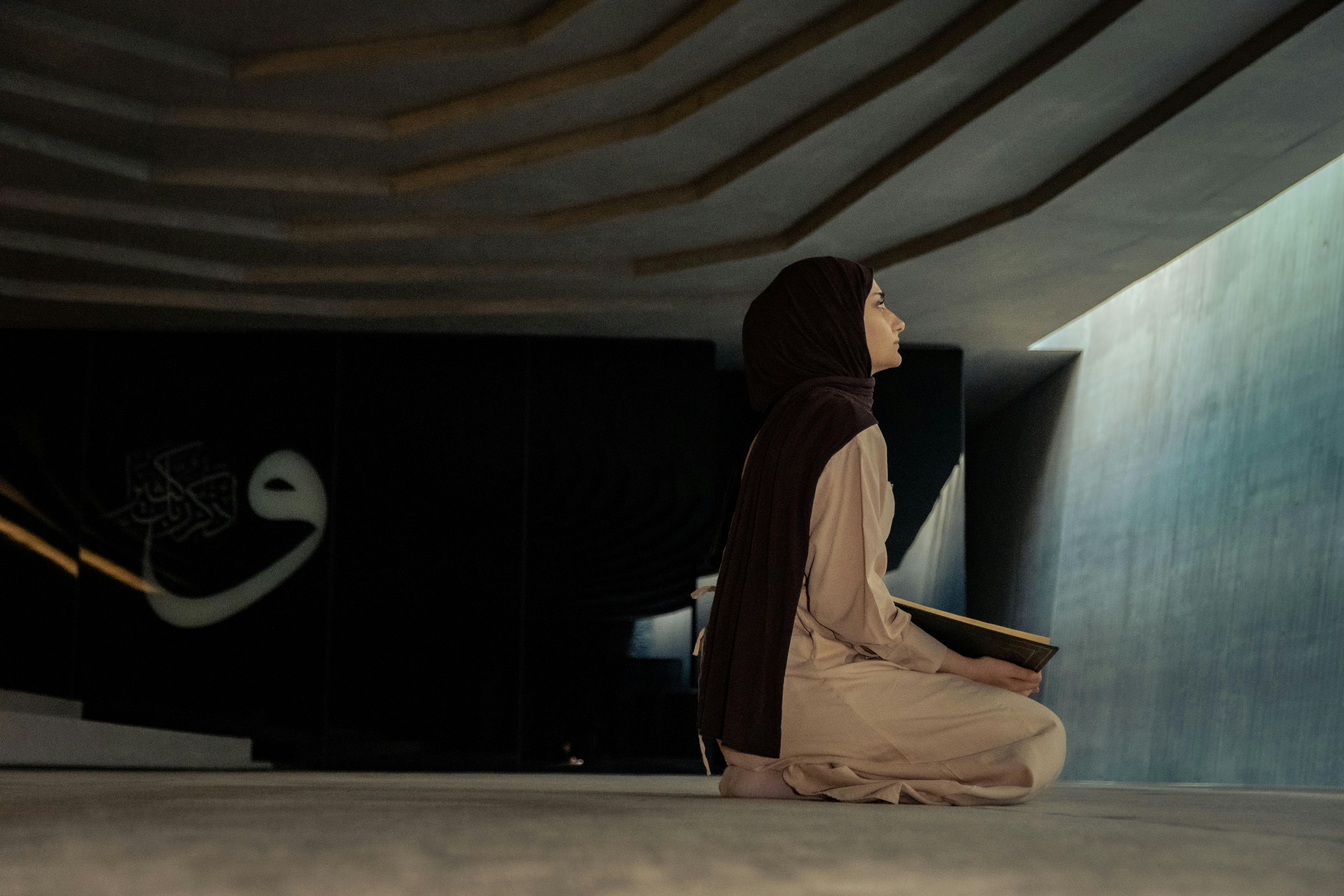 Free A woman in a hijab sits praying indoors, bathed in soft light, capturing a moment of spiritual reflection. Stock Photo