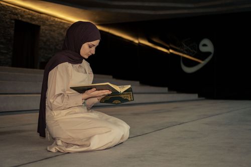 Free A woman kneeling and reading a Koran inside a dimly lit mosque, wearing a hijab. Stock Photo