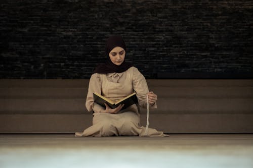 A woman in a hijab sitting on the floor reading a book