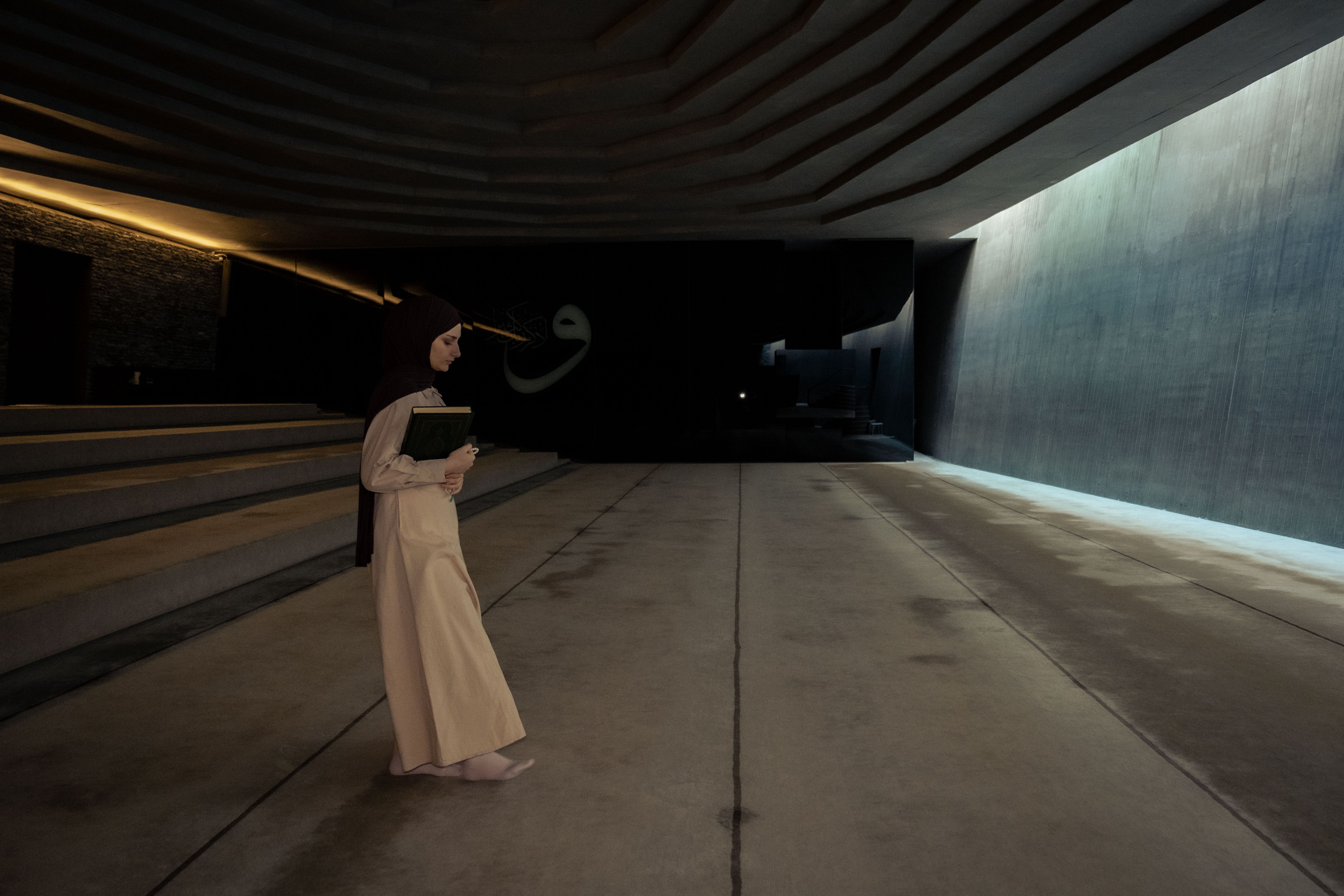 A woman in a hijab walks through a modern, dimly lit hallway holding a book, emphasizing cultural and religious themes.