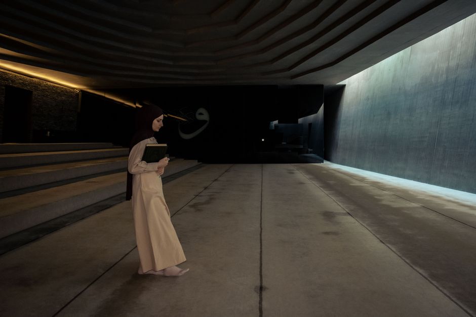 A woman in a hijab walks through a modern, dimly lit hallway holding a book, emphasizing cultural and religious themes.