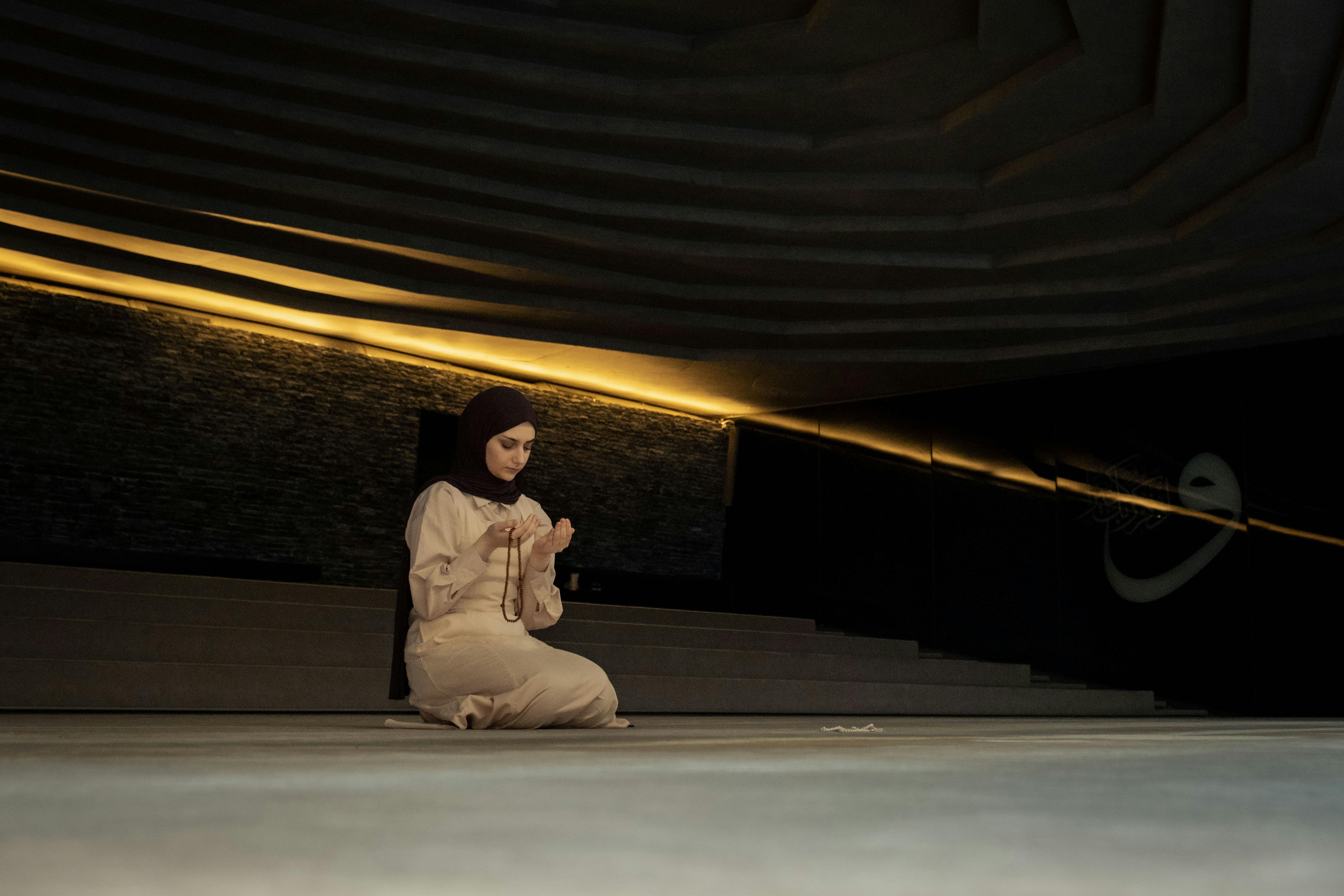 Middle Eastern woman in hijab kneeling in a serene, dimly lit space holding prayer beads.