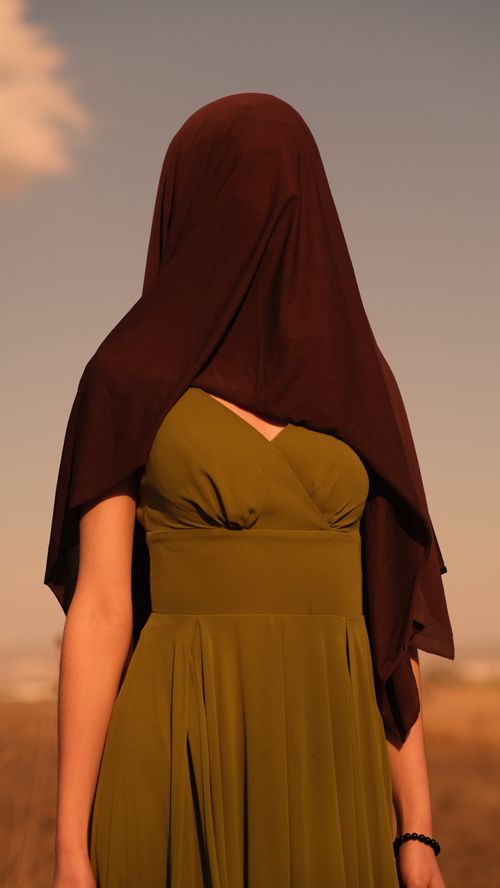 Free A woman in an olive green dress with a headscarf stands outdoors during sunset, creating an enigmatic scene. Stock Photo